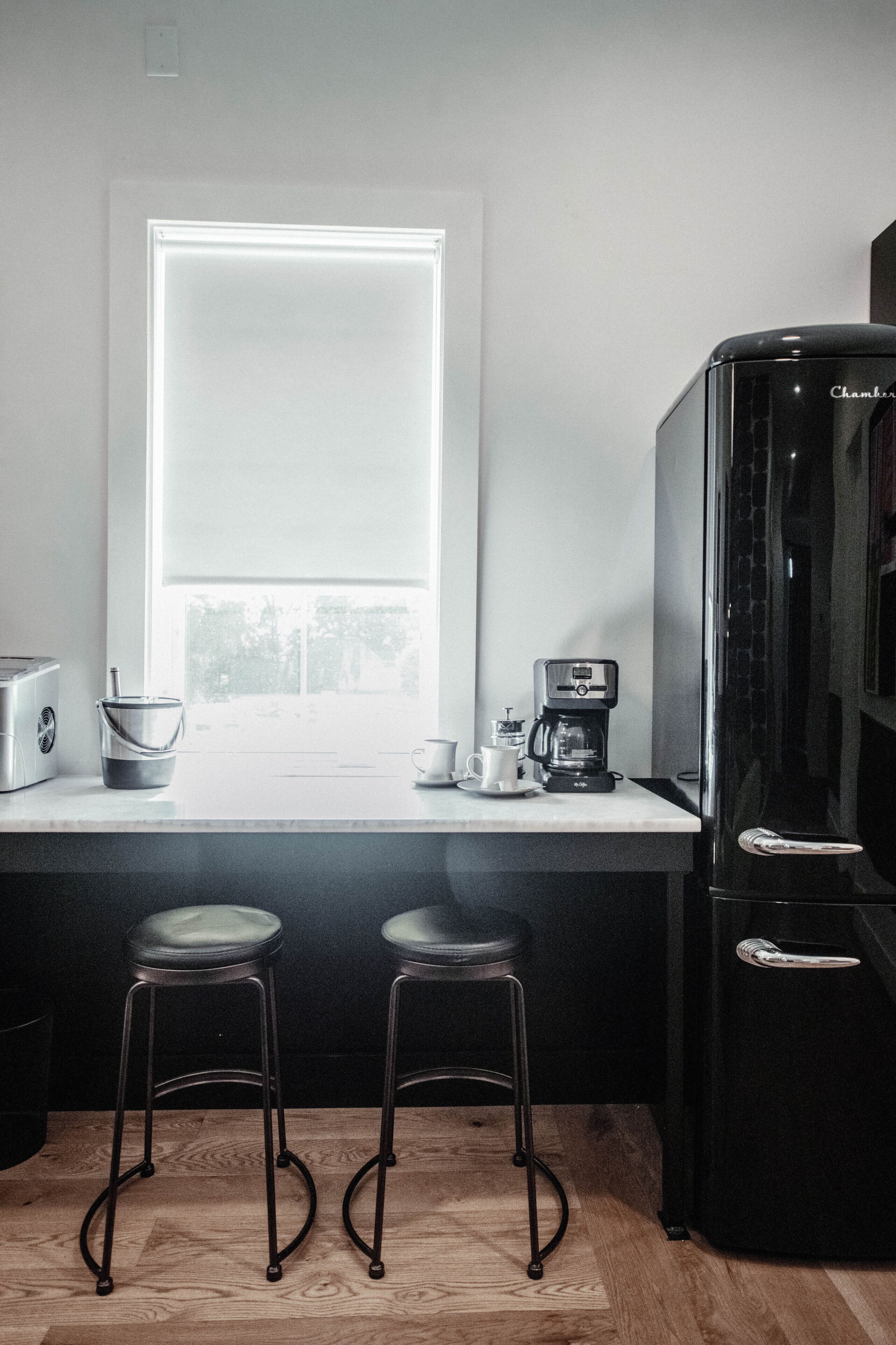 A kitchen with a window, a coffee maker, two stools, and a refrigerator.