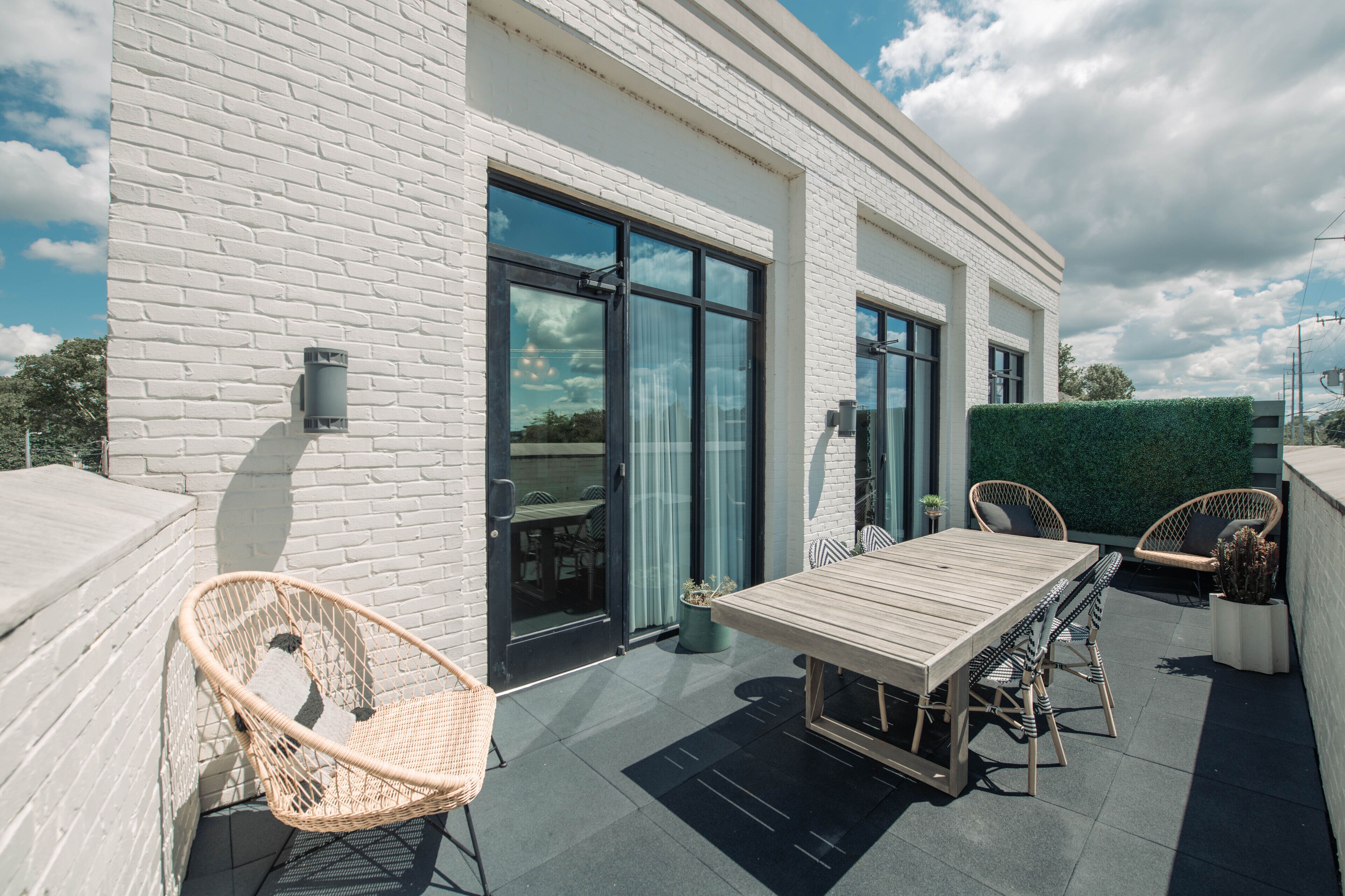 Un bâtiment moderne à l’extérieur en briques blanches, doté de grandes portes vitrées et d’un patio avec une table en bois et des chaises en osier.