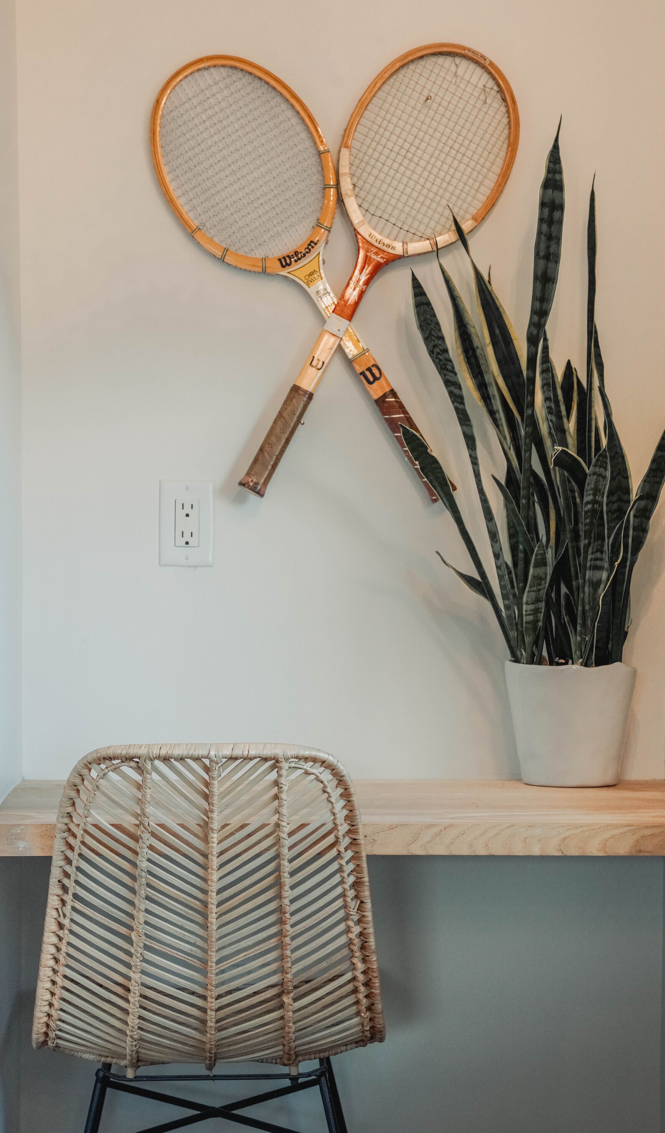 A wall-mounted tennis racket, a woven chair, and a potted plant.