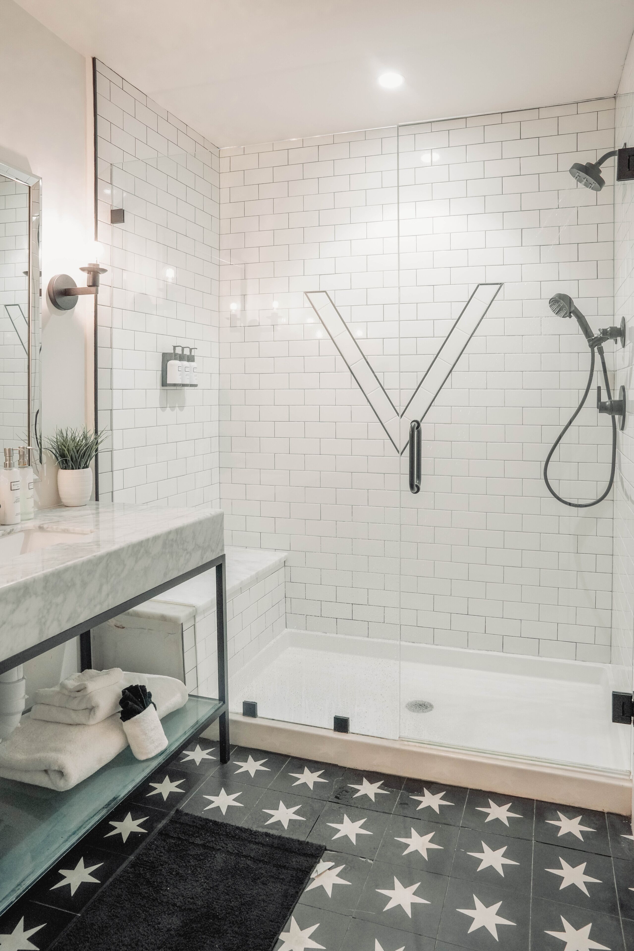 Une salle de bain moderne avec des carreaux de métro blancs, un miroir en forme de V, une douche en verre et un sol orné d’un motif en étoile.