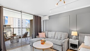 Une chambre d’hôtel moderne avec un canapé gris, une table basse ronde, une petite table d’appoint avec une lampe, et une vue sur la ville grâce à de grandes fenêtres.