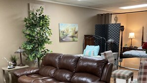 A living room with a brown leather sofa, a patterned armchair, a small side table with a lamp, a large potted plant, and a painting on the wall. - Aunt Minnie's Peabody Flat near Mountain View's Historic Courthouse Square (Mountain View)