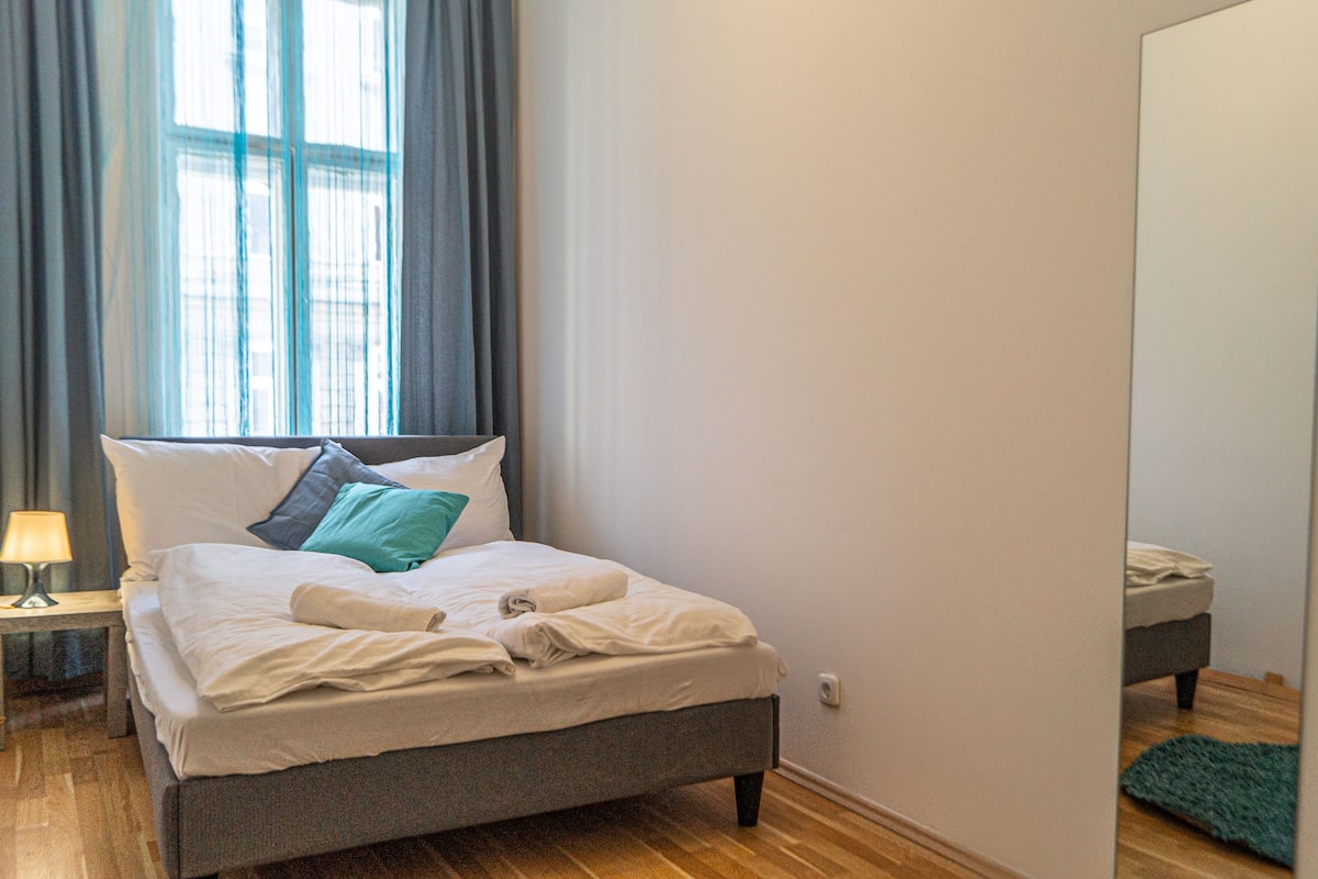 A neatly made bed with pillows, a bedside table with a lamp, and a view of a window with curtains.
