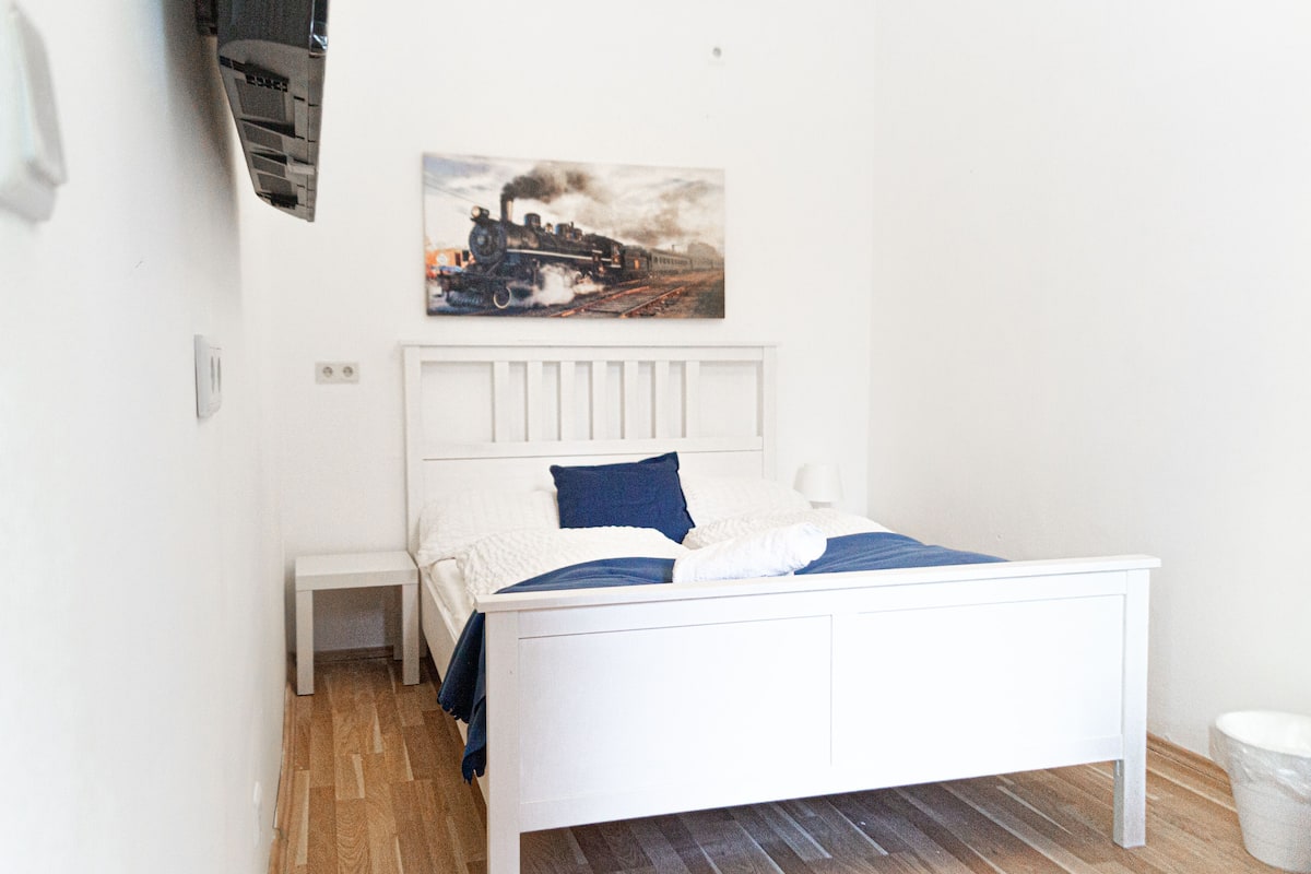 A white bed with a blue pillow and a blanket, a small bedside table, and a wall-mounted picture of a train.