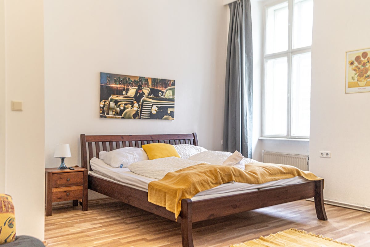 A bedroom with a wooden bed, a nightstand, a lamp, a painting on the wall, and a window with curtains.
