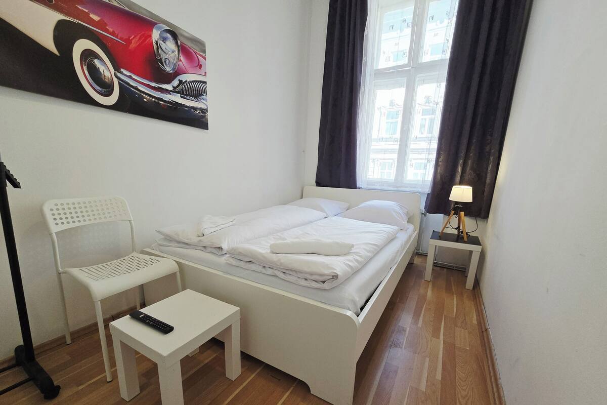 A neatly made bed with white bedding, a white chair, a small table with a remote control, a lamp on a bedside table, and a painting of a car on the wall.