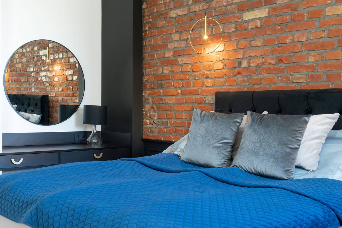 Quarto com parede de tijolos, um espelho redondo, cama com roupa de cama azul e cabeceira preta.