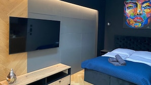 Un dormitorio moderno con una televisión de pantalla grande, un lecho con colcha azul y un cuadro abstracto colorido sobre la cama.