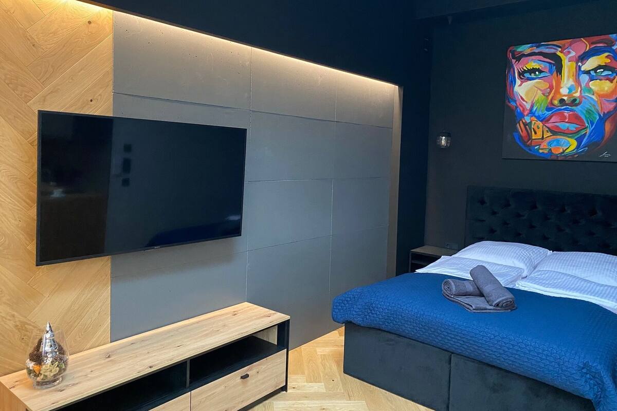 Quarto moderno com uma grande TV de tela plana fixada em uma parede revestida de madeira, uma cama com colcha azul e uma pintura abstrata colorida sobre a cama.
