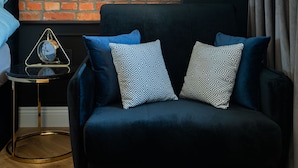 Una habitación con pared de ladrillo, un sofá azul oscuro con cojines estampados, una mesita auxiliar con un reloj y una lámpara colgante.