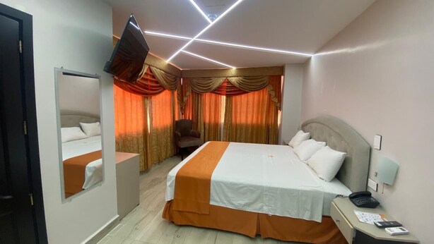 A hotel room with a large bed, a bedside table with a lamp, a TV mounted on the wall, and a window with curtains. - BRITNEY HOTEL MACHALA (Machala)