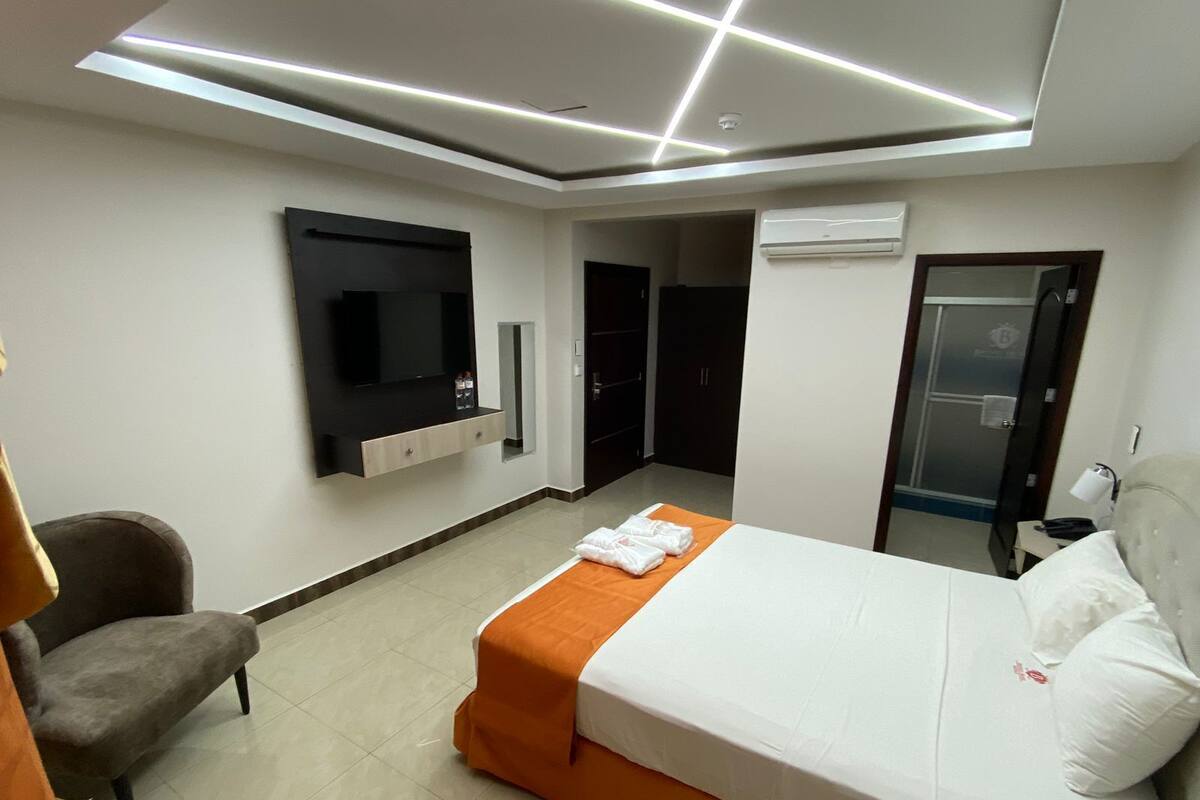 A modern hotel room with a bed, a TV mounted on the wall, a chair, and a door leading to a bathroom.