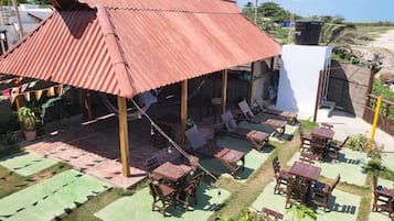 Un espace extérieur abrité, doté de tables et de chaises, entouré de verdure et surmonté d’un toit en métal ondulé.