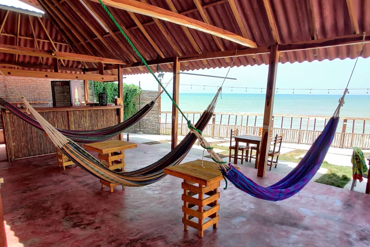 A covered outdoor area with wooden furniture, including a hammock and a small table, overlooking a beach.