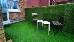 A small outdoor area with artificial grass, a bench, two white tables, and a hedge. - Apartment Sleeps 7 With Wifi (London)