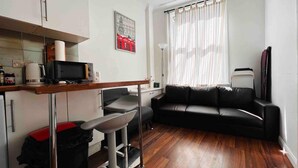 A compact living space with a kitchenette, a black leather sofa, a dining area with a bar stool, and a large window with sheer curtains. - Apartment Sleeps 7 With Wifi (London)