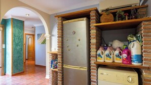 Cozinha equipada com frigorífico, máquina de lavar loiça e estantes com produtos de limpeza.
