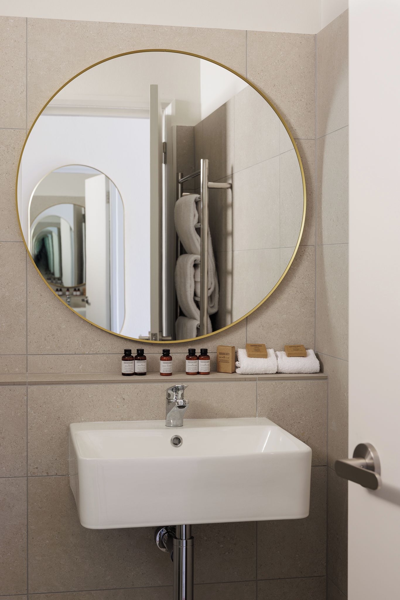 Une salle de bain avec un miroir rond, un lavabo blanc et des articles de toilette rangés sur une étagère.