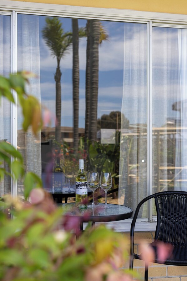 A glass table with a bottle and two glasses, a black chair, and a view of palm trees through a window with sheer curtains.