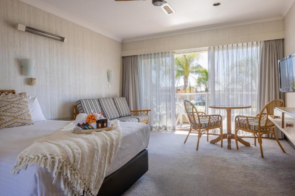 Habitación de hotel con cama, zona de comedor con sillas de mimbre, televisor y balcón con cortinas.