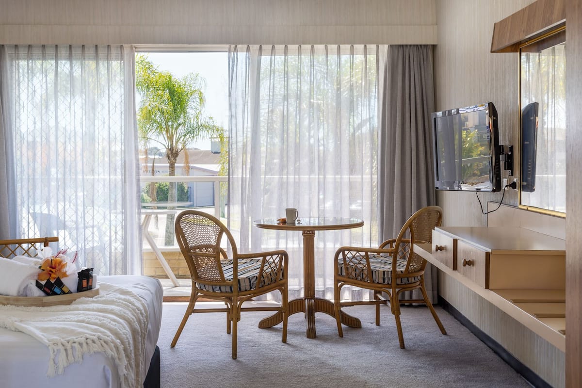 Habitación de hotel con una cama, dos sillones de mimbre, una mesita redonda, un televisor montado en la pared y una ventana con cortinas traslúcidas.