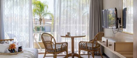 Une chambre d’hôtel avec un lit, deux fauteuils en osier, une petite table ronde, un téléviseur fixé au mur et une fenêtre avec des rideaux légers.