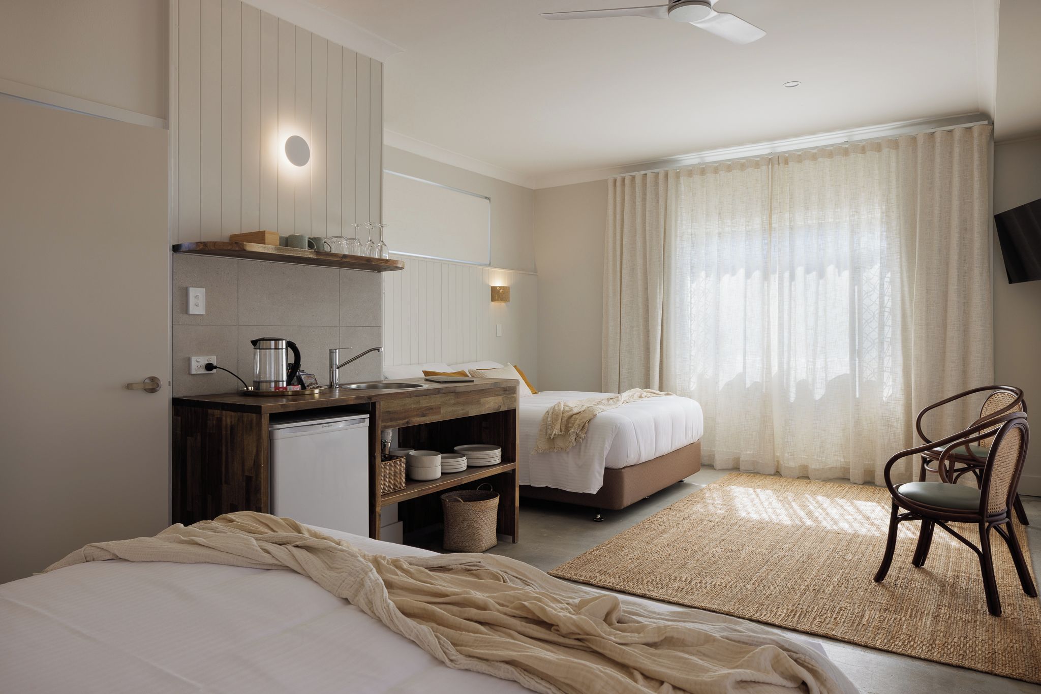 Ein Hotelzimmer mit einer Küchenzeile, zwei Betten, einem Sessel und einem großen Fenster mit Vorhängen.