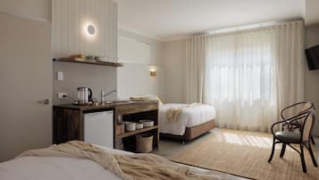A hotel room with a kitchenette, two beds, a chair, and a large window with curtains.