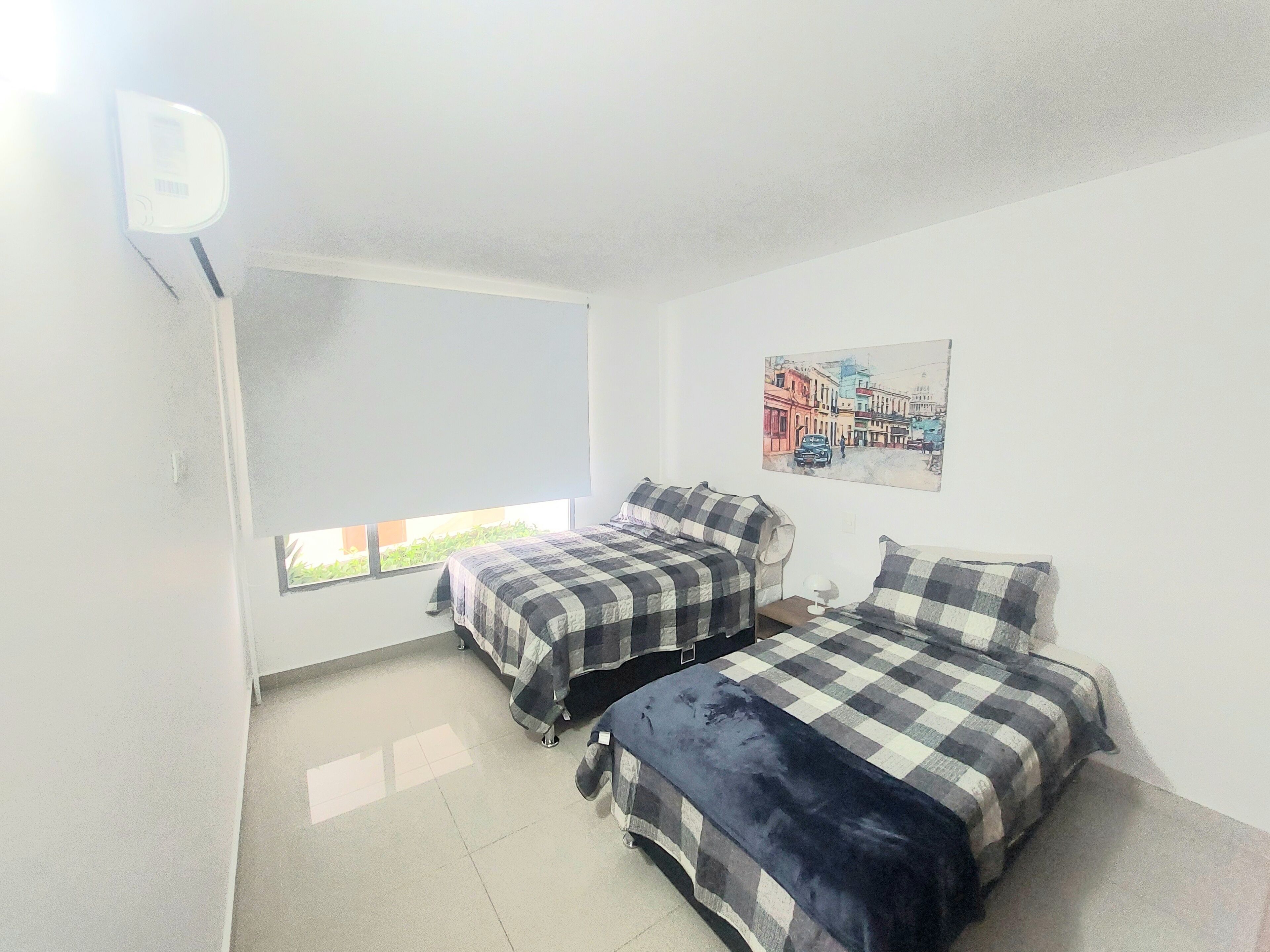 Habitación con dos camas, una ventana, aire acondicionado instalado en la pared y una pintura con vista a la ciudad.