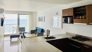 Une cuisine moderne dotée d’un grand îlot central, un coin repas offrant une vue sur la mer et un espace de vie avec un canapé et un téléviseur.