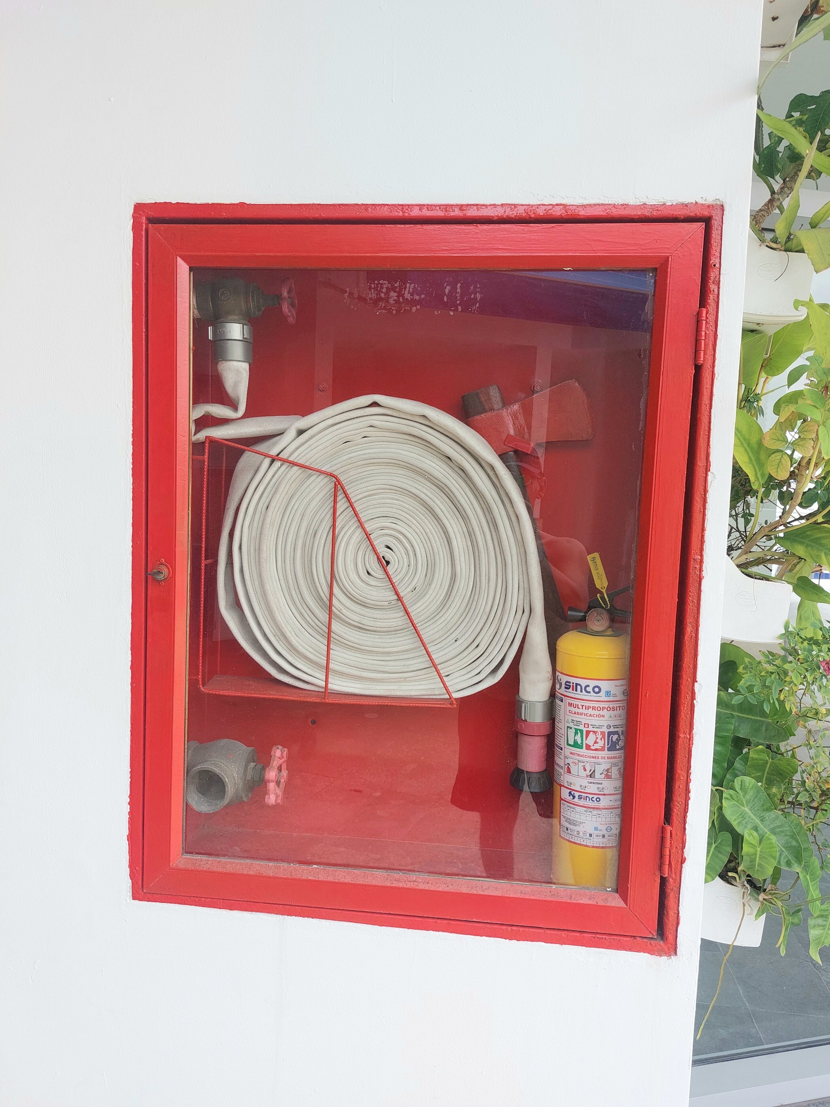 En brandsläckare som är monterad på en vägg, med en röd ram, en hoprullad vit slang och en gul brandsläckare inuti ett glasskåp.