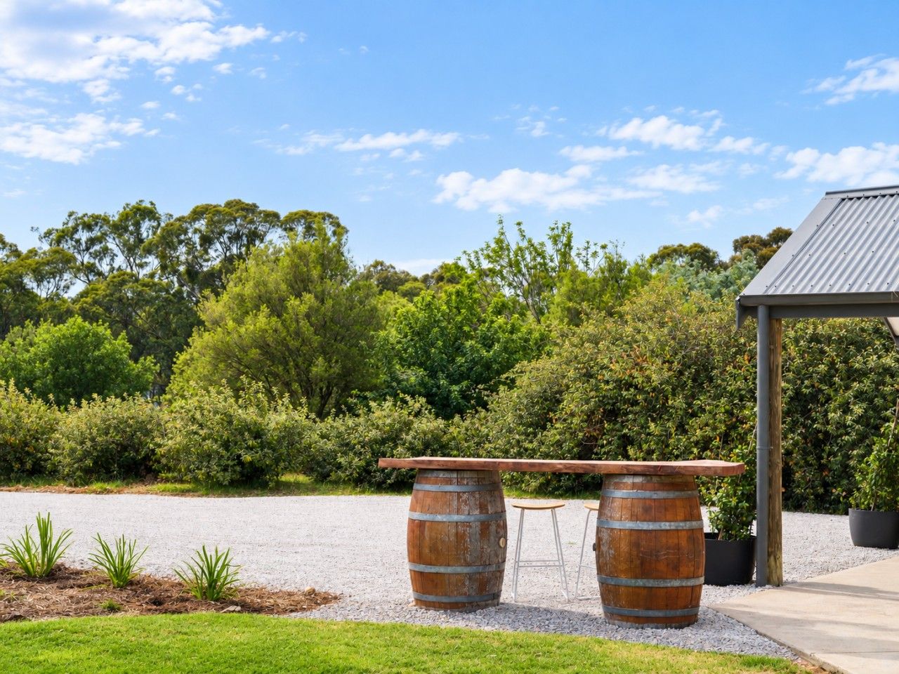 A rustic outdoor seating area with two wooden barrels and a bench, surrounded by greenery and a clear sky.