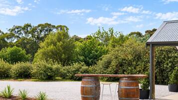 A rustic outdoor seating area with two wooden barrels and a bench, surrounded by greenery and a clear sky.