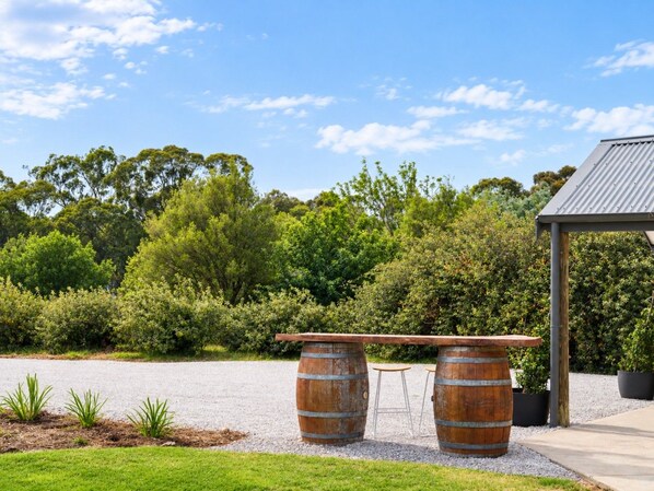 A rustic outdoor seating area with two wooden barrels and a bench, surrounded by greenery and a clear sky.