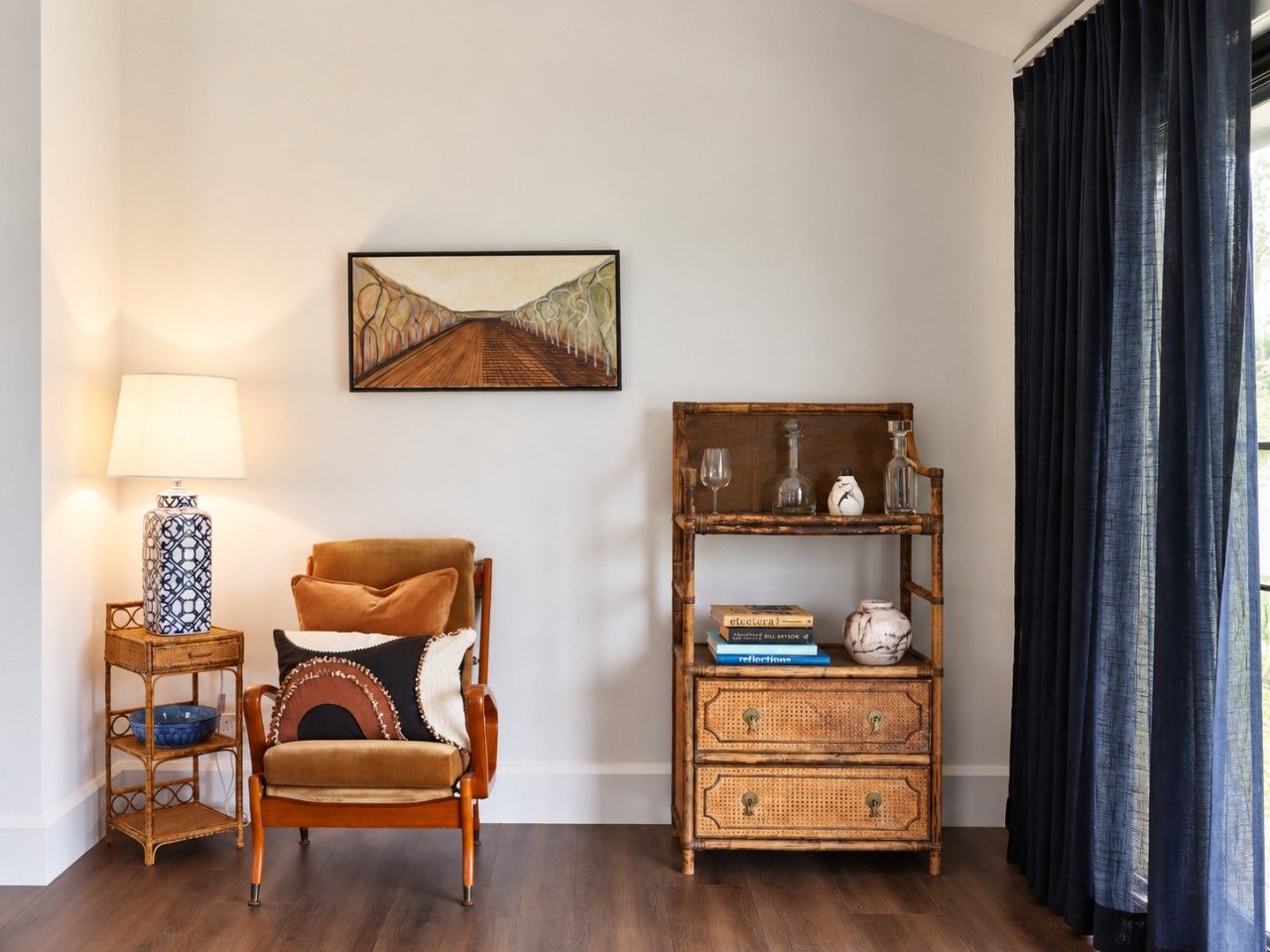 A room with a wooden chair, a side table with a lamp, a bookshelf, and a painting on the wall.