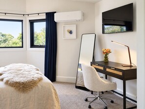 A hotel room with a bed, a desk with a chair, a large mirror, an air conditioner, a flat-screen TV, and a window with a view of trees.