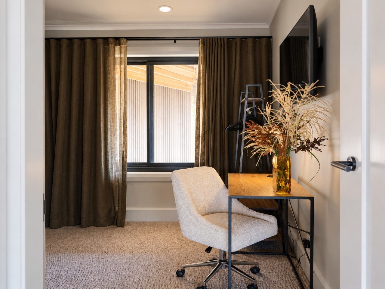A room with a desk, chair, and a vase with dried plants.