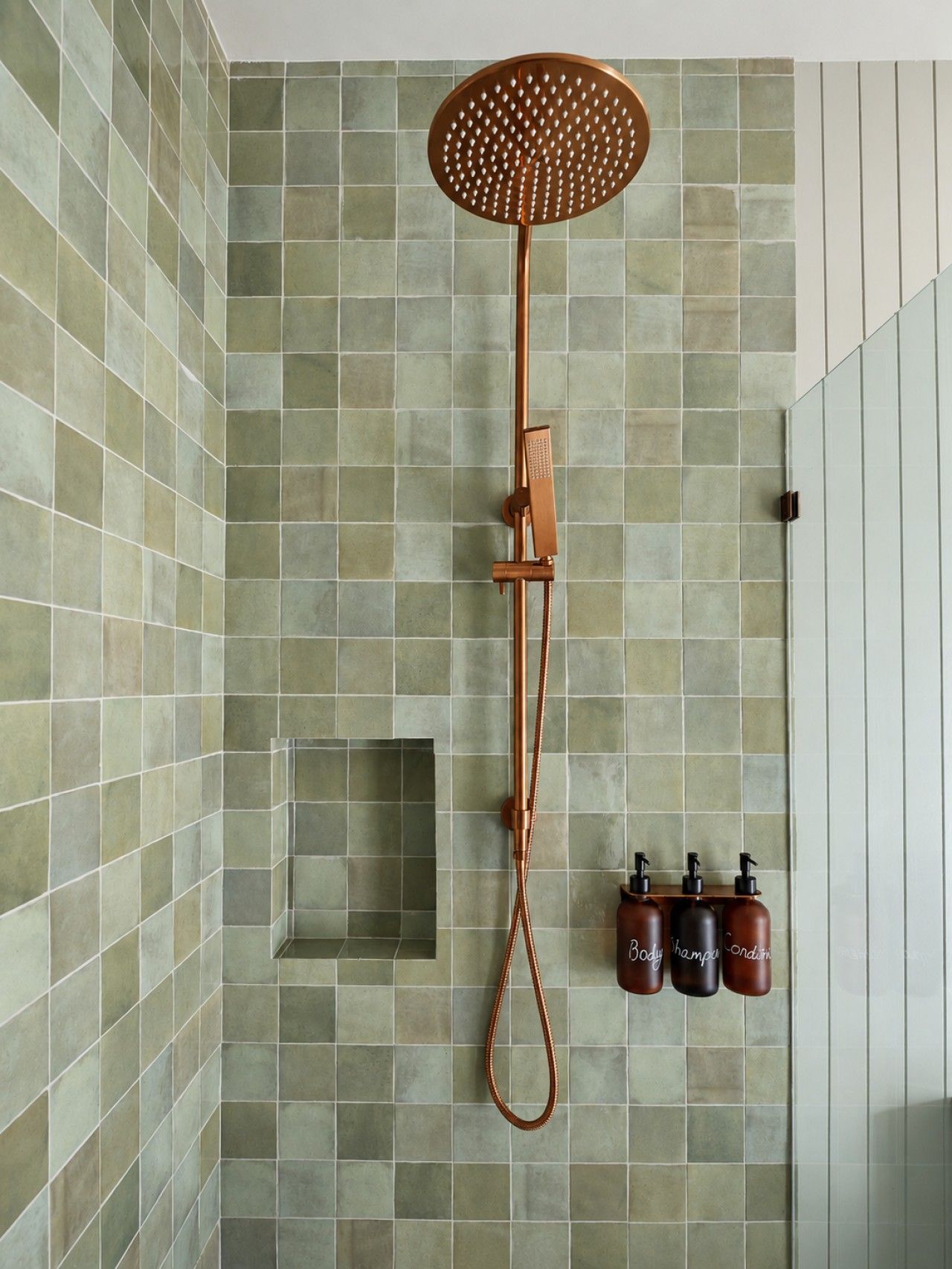 Une douche équipée d’un pommeau amovible, d’un flexible de douche et de trois bouteilles de savon placés sur une étagère fixée au mur.