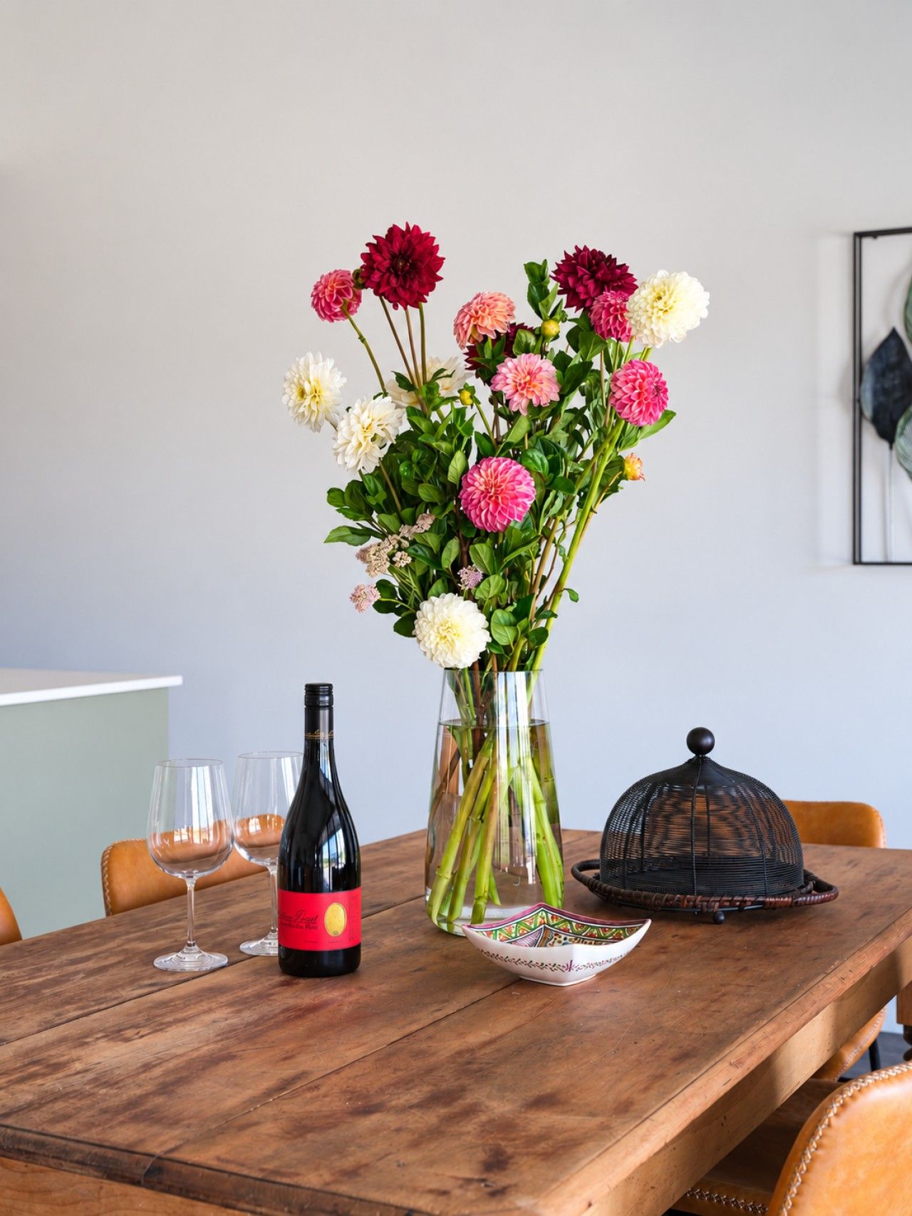 A wooden table with a wine bottle, two glasses, a vase with flowers, and a decorative bowl.