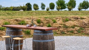 Ein rustikaler Sitzbereich im Freien mit einem Holztisch und Hockern, vor einer grünen Wiese und Bäumen gelegen.