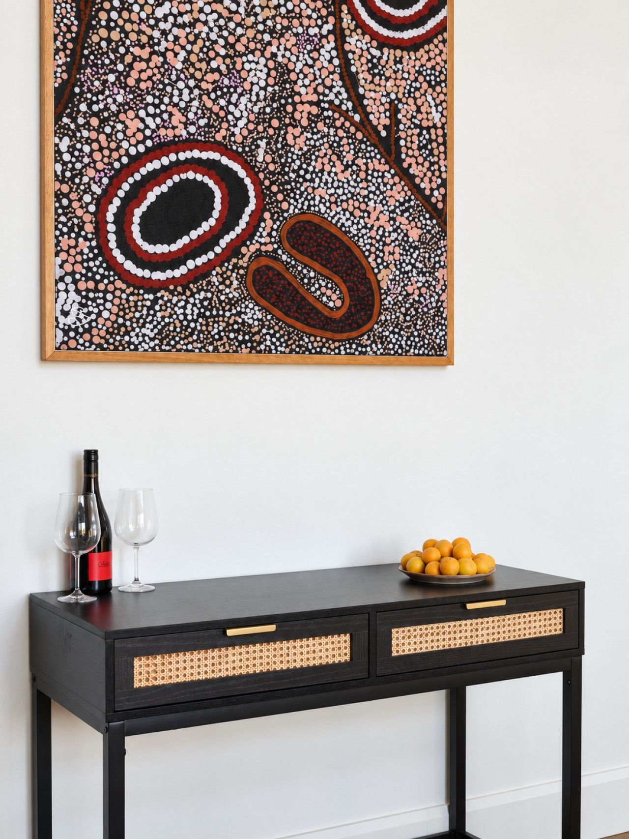 A black console table with woven side panels, a wine bottle, two wine glasses, and a bowl of oranges against a white wall.