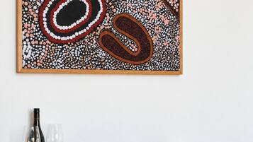 A black console table with woven side panels, a wine bottle, two wine glasses, and a bowl of oranges against a white wall.