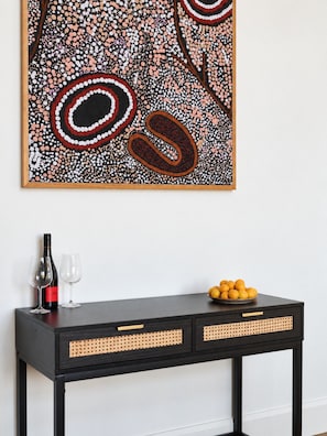 A black console table with woven side panels, a wine bottle, two wine glasses, and a bowl of oranges against a white wall. - Shawwood Bottling Room - Luxury Mudgee Farm Stay, close to town and wineries (Bombira)