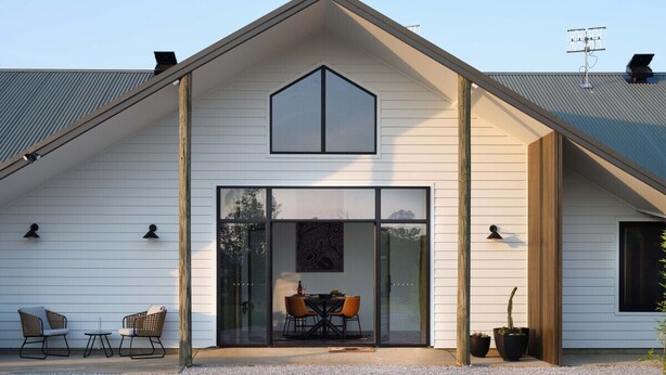 A modern house with a large sliding glass door entrance, a dining area with two chairs and a table, and a small garden with potted plants.