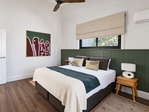 Un dormitorio con cama, mesita de noche, cuadro en la pared, ventana con persiana y ventilador de techo.