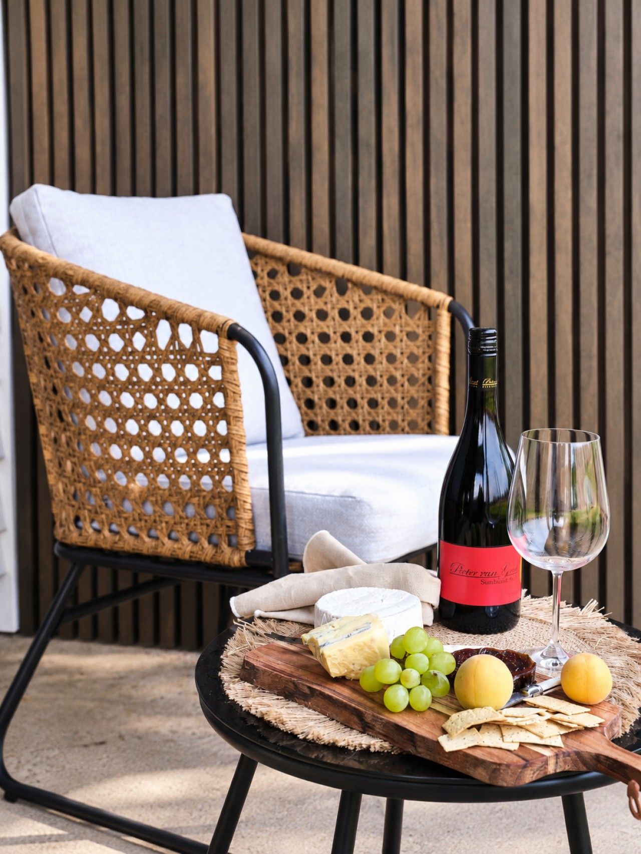 Une chaise en osier avec un coussin blanc, une bouteille de vin, un verre et une planche en bois sur laquelle sont disposés du fromage, des raisins et des crackers.