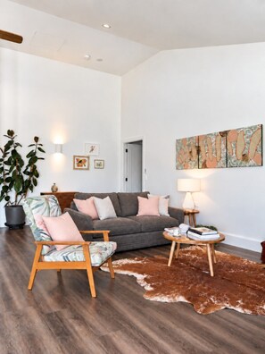 Un salón amplio con un sofá gris, una silla de madera, una mesa redonda de centro y un cuadro abstracto grande en la pared.