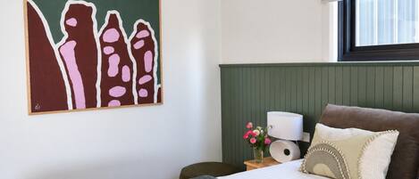 A bedroom with a bed, a nightstand, a lamp, a painting, and a window with a blind.