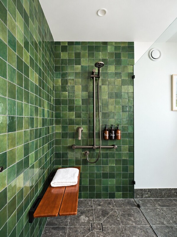A bathroom with green-tiled walls, a wooden bench, a shower with a handheld showerhead, and a towel rack.