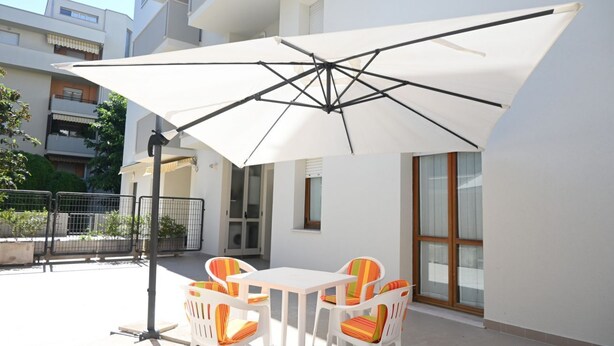 Un patio al aire libre con una mesa y sillas blancas, un paraguas grande de color blanco y vista a un edificio residencial al fondo.